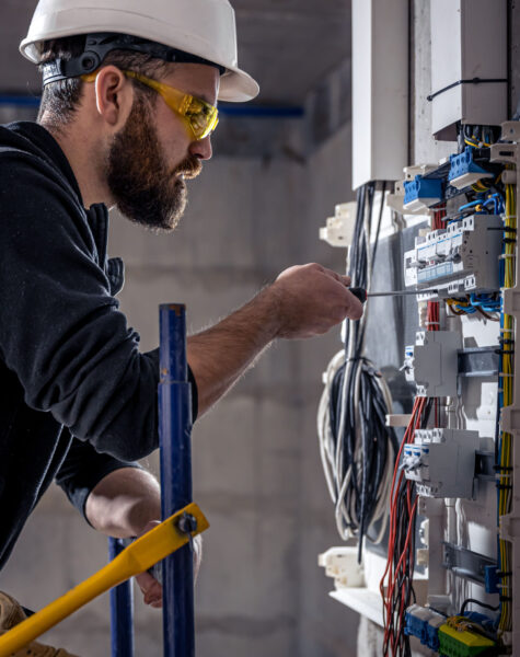 A male electrician works in a switchboard with an electrical connecting cable, connects the equipment with tools.